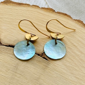 May include: A pair of gold-colored drop earrings. Each earring features a small, half-moon shaped accent above a round, iridescent, light blue disc. The earrings are displayed on a wooden surface.
