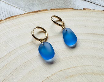 May include: A pair of gold hoop earrings with teardrop-shaped, frosted blue glass pendants. The earrings are displayed on a light-colored wooden surface, showcasing the jewelry's design and color.