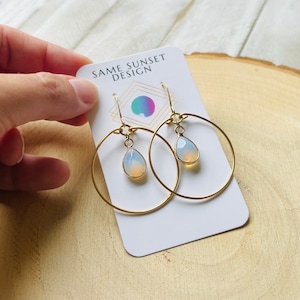 May include: Gold hoop earrings with a teardrop-shaped opal pendant. The earrings are on a white card with the text "SAME SUNSET DESIGN".