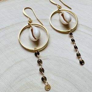 May include: Gold-colored hoop earrings with a dangling chain of small, flat, gold-colored discs. Each earring features a white seashell suspended within the hoop. The earrings are displayed on a light-colored wooden surface.