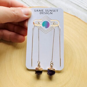 Puede incluir: Pendientes colgantes dorados con piedras de amatista en bruto. Los pendientes tienen una tapa dorada en las piedras de amatista. Se muestran en una tarjeta blanca con el texto "SAME SUNSET DESIGN".