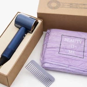 May include: A navy blue hair dryer with gold accents is displayed in a cardboard box. A gray comb and a package of lavender towels with the text "BEAUTY -II- ME" are also visible. The hair dryer is a modern design.