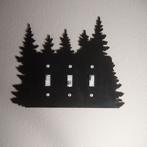 Puede incluir: Tapa de interruptor de luz de metal negro con una silueta de un bosque de pinos. La tapa tiene tres interruptores.