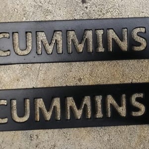 May include: Two black metal plates with the word "CUMMINS" cut out. Each plate has a rectangular shape with rounded corners and a hole on each end. The plates are lying on a textured gray surface.