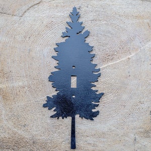 Op de afbeelding: Zwarte metalen lichtschakelaar in de vorm van een dennenboom silhouet. De afdekking heeft een enkele schakelaar opening en een klein gat aan de onderkant voor montage.