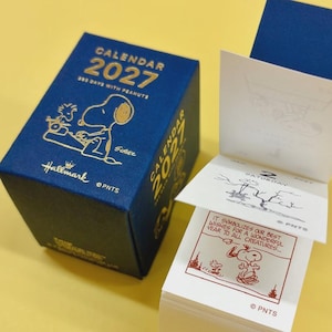 Peut inclure: Une boîte bleue pour un calendrier quotidien 2027 avec du texte doré et une illustration de Snoopy, à côté d'une pile de pages de calendrier blanches détachables. La page supérieure affiche une bande dessinée avec le texte, It symbolizes our best wishes for a wonderful year to all creatures.