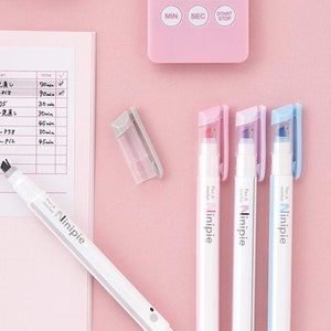 May include: A white pen and highlighter combo with the word "Ninipie" printed on the side. The pen is on a pink surface with a notebook and other stationery items.