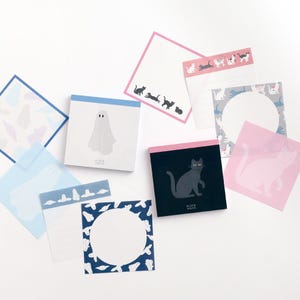 May include: A selection of square and rectangular notepads with diverse designs. Some pads feature ghost and cat illustrations in black and white, while others have patterned borders. The pads are in shades of blue, pink, and white.