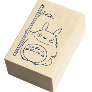May include: A wooden stamp with a blue line drawing of a cartoon character holding a stick. The character has large eyes and a wide smile. The character is surrounded by a light brown background.