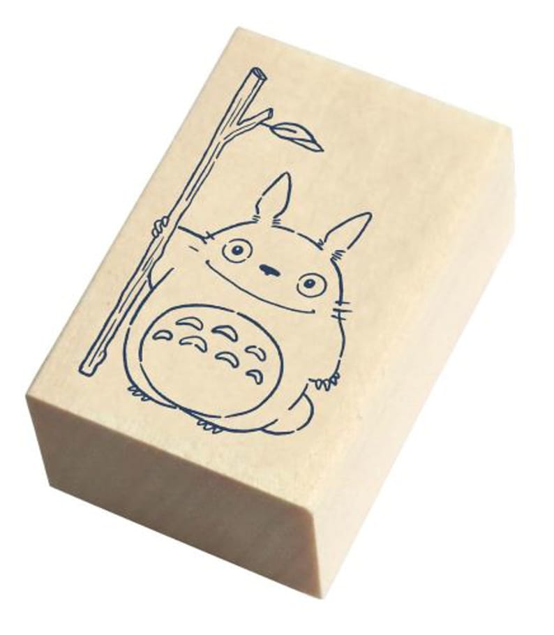 May include: A wooden stamp with a blue line drawing of a cartoon character holding a stick. The character has large eyes and a wide smile. The character is surrounded by a light brown background.