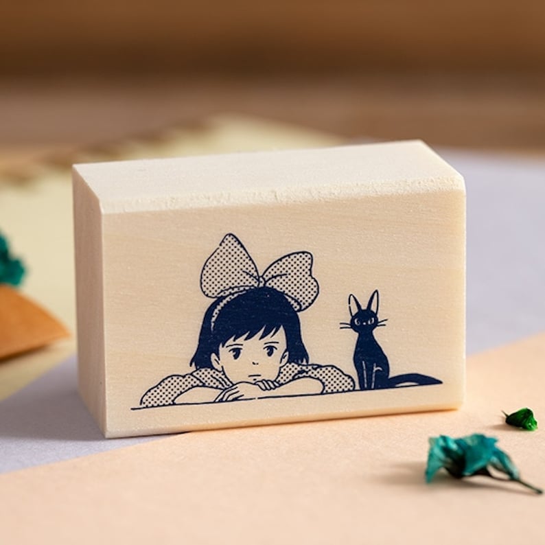 May include: A wooden stamp with a black and white illustration of a girl with a bow in her hair and a black cat. The girl is looking to the side and the cat is looking at the girl.