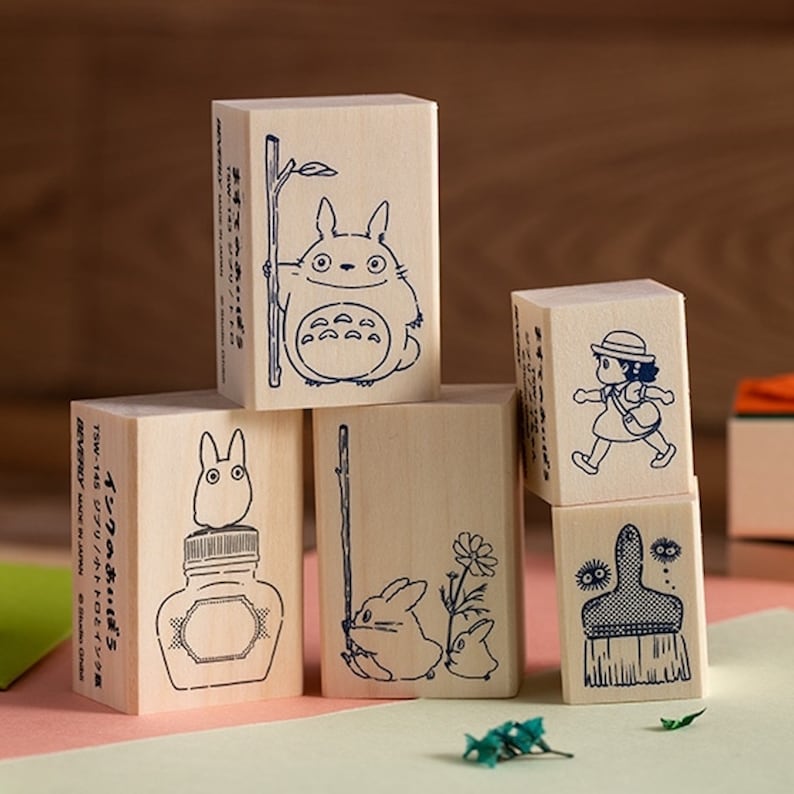 May include: Set of five wooden stamps featuring characters from the animated film 'My Neighbor Totoro'. The stamps are rectangular and have a light wood finish. The designs include Totoro, a small Totoro, a girl with a hat, a broom, and a bottle of ink. The text on one of the stamps reads 'Ink's Friend' in Japanese.