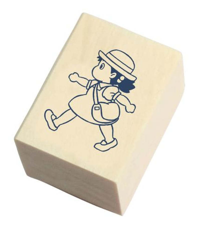 May include: A wooden rubber stamp with a blue line drawing of a girl wearing a hat and carrying a bag. The girl is walking to the right.
