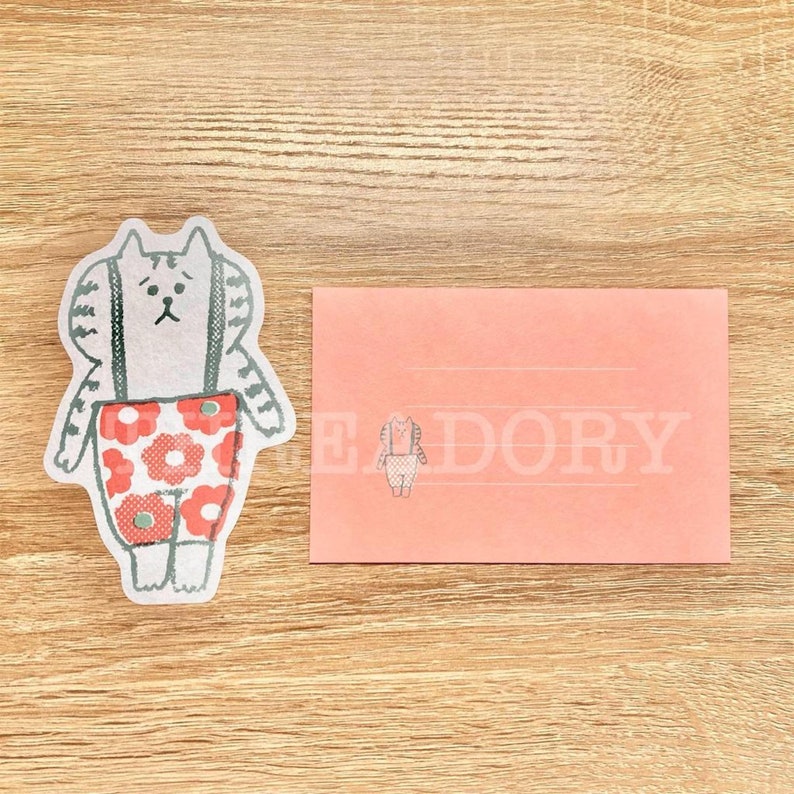 Może przedstawiać: R&oacute;żowa koperta z ilustracją białego kota w czerwonych spodniach z kwiatowym wzorem. Kot ma na sobie szelki i smutny wyraz twarzy. Koperta ma wyściełane wnętrze i napis "LEDORY" nadrukowany z przodu.