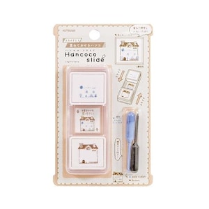 Puede incluir: Un juego de sellos Hancoco slide con tres sellos cuadrados con ilustraciones de casas. El juego incluye un aplicador de tinta azul y negro. El texto en el paquete dice "Hancoco slide", "pale violet" y "brown".