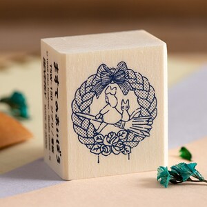 May include: A wooden stamp with a blue design of a witch riding a broom with a cat. The witch is wearing a hat and a bow. The design is surrounded by a wreath with a bow. The text on the stamp reads "まほうつかいとねこ" in Japanese.