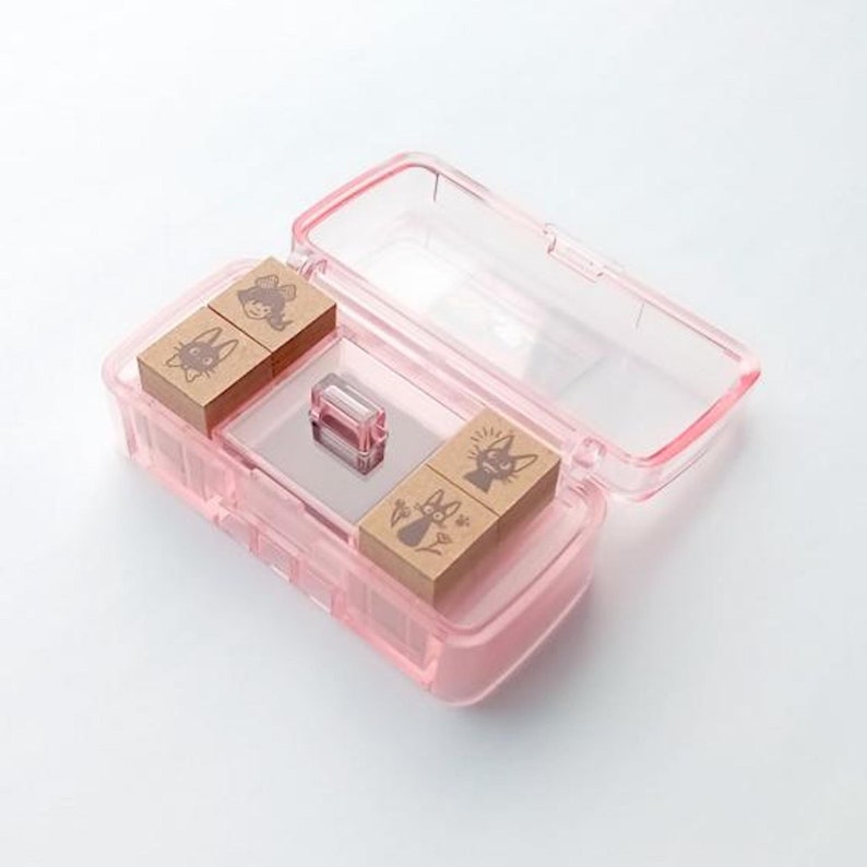 May include: A set of four small, wooden stamps in a clear pink plastic case. The stamps feature illustrations of a cat with a star above its head, a cat with flowers, a cat with a crescent moon, and a girl with a cat on her head.