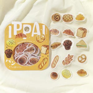 May include: A collection of 45-piece deco stickers depicting baked items such as croissants, bread, and muffins. The stickers are presented in a box with the word "IPPAI" and Japanese text. The stickers are on a white background.