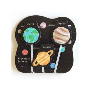 May include: A black and white card with a space theme. The card has three planets on it: Earth, Jupiter, and Saturn. The planets are drawn in color and have their names written above them. The card also has the words "Planetary Science" written on it.