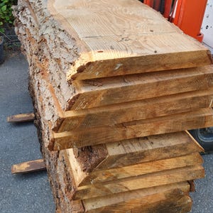 May include: A stack of large, rough-cut wooden slabs with bark still attached. The slabs are light brown in color and have a natural, rustic look.
