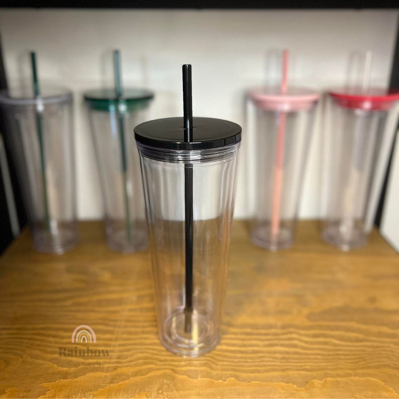 以下が含まれることがあります： 黒い蓋と黒いストローが付いた透明なプラスチック製のタンブラー。タンブラーは空で、木製の表面の上に置かれています。