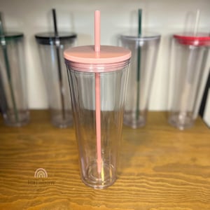 以下が含まれることがあります： ピンクの蓋とピンクのストローが付いた透明なプラスチック製のタンブラー。タンブラーは空で、木製の表面の上に置かれています