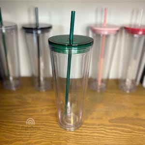 以下が含まれることがあります： 緑色の蓋とストローが付いた透明なプラスチック製のタンブラー。タンブラーは木製の表面の上に置かれています。