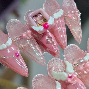 Könnte beinhalten: Rosa Acrylnägel mit verschiedenen Verzierungen. Einige Nägel haben weiße Engelsflügel, rosa Schleifen und kleine Perlen. Ein Nagel hat ein kleines gerahmtes Bild einer Person. Die Nägel sind hellrosa mit glänzendem Finish.