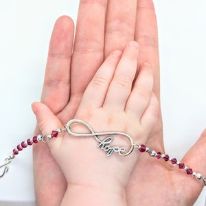 May include: A silver infinity bracelet with the word "hope" and red beads. The bracelet is held over a child's hand, resting on an adult's palm. The beads are a deep red color, and the bracelet's clasp is visible.