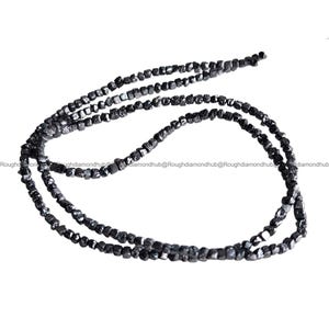 May include: Strands of black rough-cut diamond beads. The beads are irregularly shaped and have a matte finish. The strands are draped against a white background, showcasing the dark color and texture of the diamonds. The image is well-lit, highlighting the details of the gemstones.