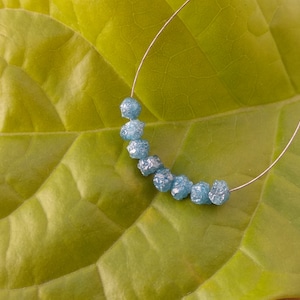 Könnte beinhalten: Eine Strähne aus hellblauen, facettierten, rohen Edelsteinperlen auf einem silbernen Draht. Die Perlen sind in einem Halbkreis auf einem grünen Blatt angeordnet.
