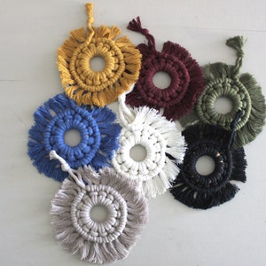 Può includere: Sette arazzi macramé fatti a mano in vari colori, tra cui giallo, blu, bianco, nero, verde e bordeaux. Ogni arazzo presenta un design circolare con un bordo a frangia.