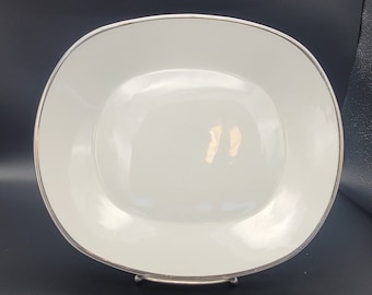 Puede incluir: Plato de servir ovalado blanco con borde plateado. El plato está sobre un soporte plateado con tres patas.