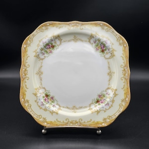 May include: A square, white porcelain plate with a gold rim and floral pattern. The plate has a scalloped edge and is decorated with delicate floral designs in shades of pink, yellow, and green.