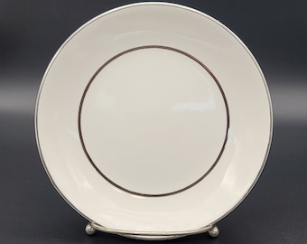 Puede incluir: Un plato de cerámica blanca con borde plateado y anillo interior marrón. El plato se exhibe sobre un soporte plateado sobre un fondo oscuro. Este elegante plato es adecuado para servir aperitivos o postres.
