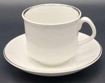 Puede incluir: Un juego de taza y platillo de porcelana blanca con borde plateado. La taza tiene un asa redondeada y el platillo tiene un borde ligeramente elevado.