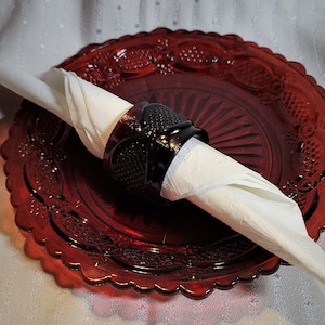 May include: A white napkin is held in place by a black glass napkin ring on a red glass plate with a scalloped edge.