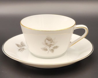 May include: A white teacup and saucer set with a gold rim and a gray rose design.