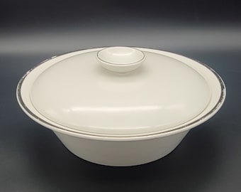 Puede incluir: Tazón de servir de cerámica blanca con tapa. El tazón tiene un borde plateado y la tapa tiene un pequeño pomo en la parte superior.