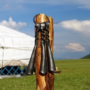 May include: Three black metal bells hanging from a wooden post. The bells are twisted and have a pointed shape. The post is brown and has a rough texture. The bells are hanging in front of a white yurt and a green grassy field.