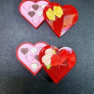 May include: Two heart-shaped boxes, one with chocolates and the other with a bow and flowers. The chocolates are in a pink box with a red border. The other box is red with a gold bow and a red bow with flowers.