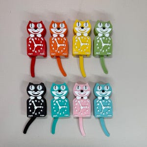 May include: A collection of eight cat-shaped wall clocks in various colors. Each clock features a cat face with a bow tie and a pendulum tail. The clocks are red, orange, yellow, green, black, teal, pink, and blue.