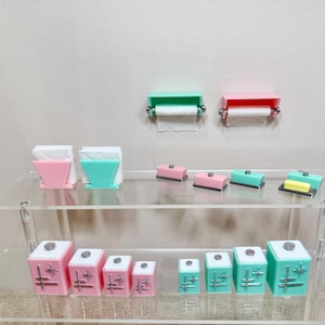 May include: A collection of miniature dolls' house accessories including two wall-mounted toilet paper holders in pink and mint green, two napkin holders, four butter dishes, and eight square canisters in pink and mint green with starburst designs.