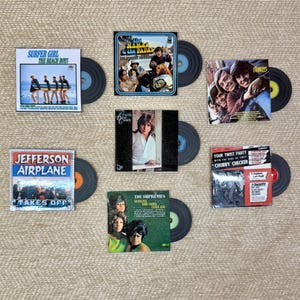 May include: A selection of vintage vinyl records with album covers. The covers display text and images, including "Surfer Girl," "The Beach Boys," "Jefferson Airplane," and "Chubby Checker." The records are arranged on a neutral surface.