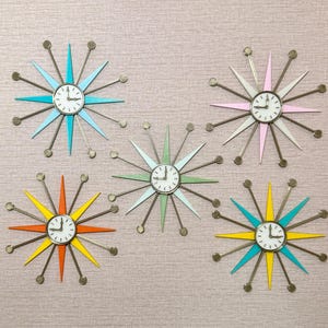 May include: Five starburst wall clocks arranged in a group. Each clock features a central white circular face with black numerals and hands, surrounded by alternating long and short metal spokes tipped with small spheres in various colors.