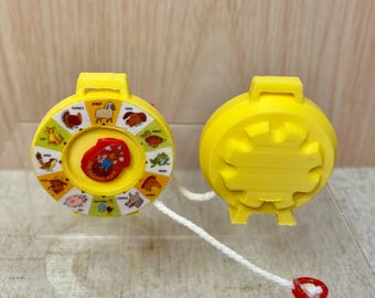 May include: A yellow plastic toy with a pull string and a spinning wheel featuring animal illustrations. The wheel has the words "cat", "bird", "pig", "chicken", "cow", "horse", "duck", "frog", "sheep", "dog", and "turkey" printed around the edge.