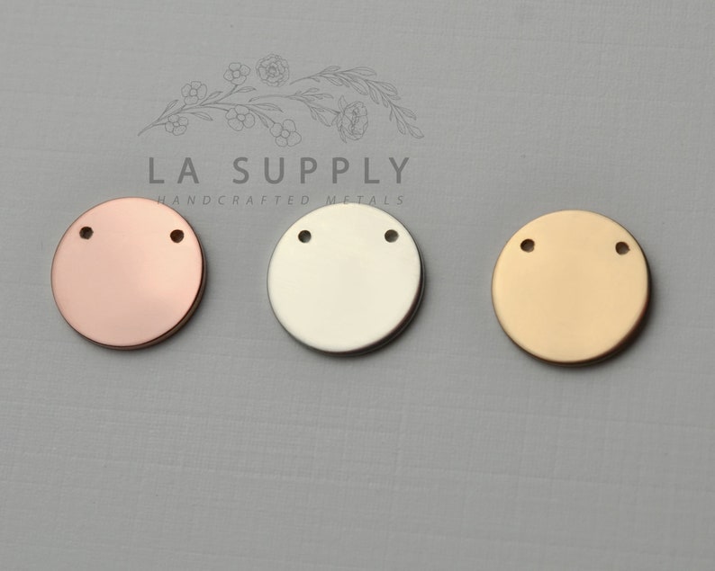 Op de afbeelding: Drie ronde metalen schijven met elk twee gaten. De schijven zijn in ros&eacute; goud, zilver en goud.