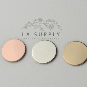 Op de afbeelding: Drie ronde metalen schijven in verschillende kleuren: ros&eacute; goud, zilver en goud. De schijven zijn in een rij van links naar rechts gerangschikt. De tekst "LA SUPPLY HANDCRAFTED METALS" staat boven de schijven.