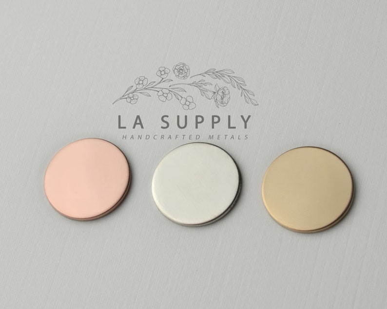 Op de afbeelding: Drie ronde metalen schijven in verschillende kleuren: ros&eacute; goud, zilver en goud. De schijven zijn in een rij van links naar rechts gerangschikt. De tekst "LA SUPPLY HANDCRAFTED METALS" staat boven de schijven.