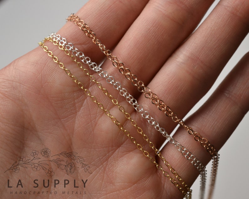 May include: Three different chain necklaces in gold, silver, and rose gold. The chains are laid out on a hand, with the gold chain on top, followed by the silver chain, and then the rose gold chain.